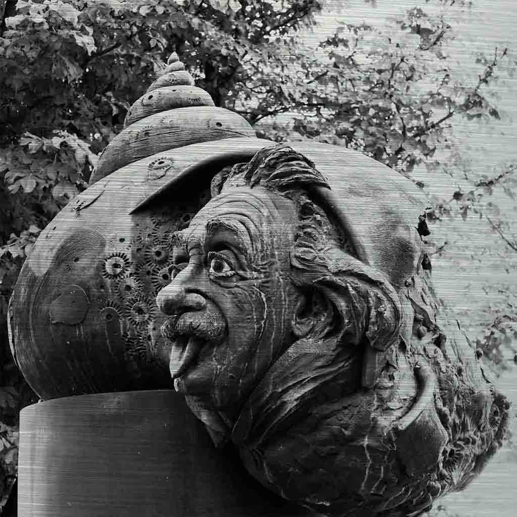 Ulm Einsteinbrunnen | Alu Dibond | #1050438 3 - 3 Ulmer Brunnenbild. Es zeigt das Bild einer Bronzeguss-Plastik von Albert Einstein. Die Seitenansicht seines Gesichts schaut mit herausgestreckter Zunge aus einem Schneckenhaus. Das Schneckenhaus wiederum liegt auf einem Raketenrumpf. Das Bild ist in den Farben Schwarz-Weiss.