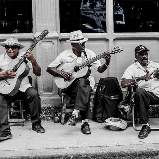 Havanna Musiker | Alu Dibond | #1080058 3 - 3 Havanna Musiker. Es zeigt das Bild von 3 dunkelhäutigen Männern, die vor einem Haus auf dem Gehweg mit ihren Stühlen sitzen und Musik machen. Zwei Männer spielen Gitarre und der andere Mann hat ein kleines Musikinstrument in den Händen. Zwei Männer tragen Hüte und ein Mann eine Baseballmütze. Alle 3 Männer sind adrett gekleidet mit einem weißen Hemd und einer dunklen Anzugshose. Im Hintergrund ist ein Haus mit Holzverkleidung und Fenstern. Das Bild ist in den Farben Schwarz-Weiß kombiniert mit Brauntönen. Das Alu Dibond Bild ist im Querformat